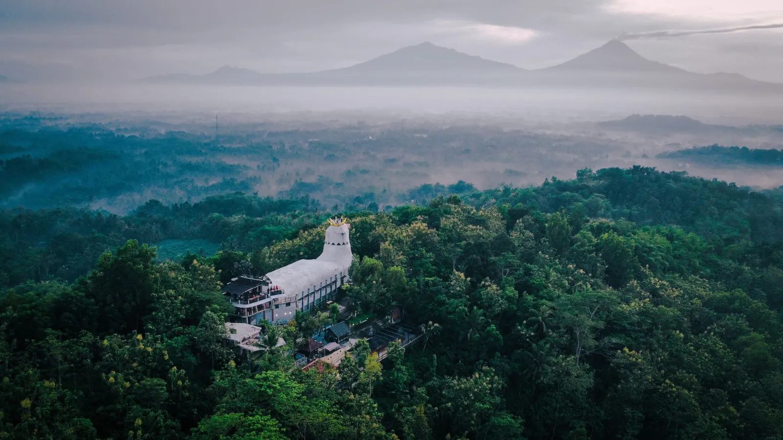 Rumah Doa Bukit Rhema berbentuk merpati bermahkota di tengah hutan Magelang dengan kabut lembah dan siluet gunung di kejauhan